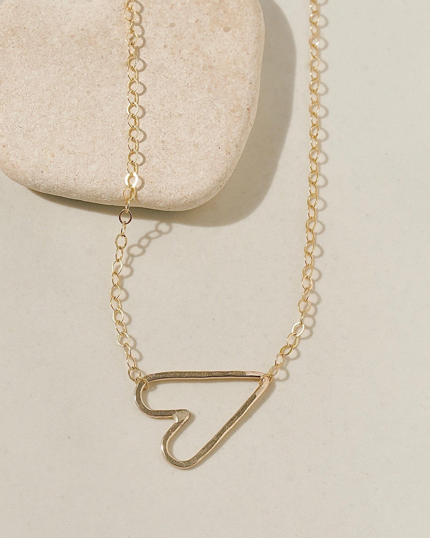 Delicate gold chain necklace with a sideways open-outline gold heart pendant on cream surface with stone pebble