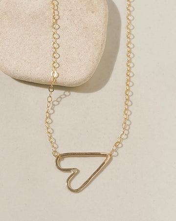 Delicate gold chain necklace with a sideways open-outline gold heart pendant on cream surface with stone pebble