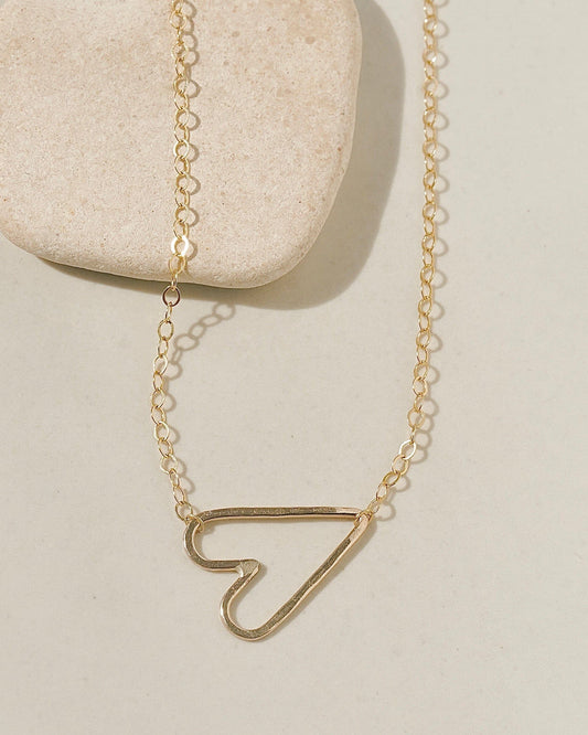 Delicate gold chain necklace with a sideways open-outline gold heart pendant on cream surface with stone pebble