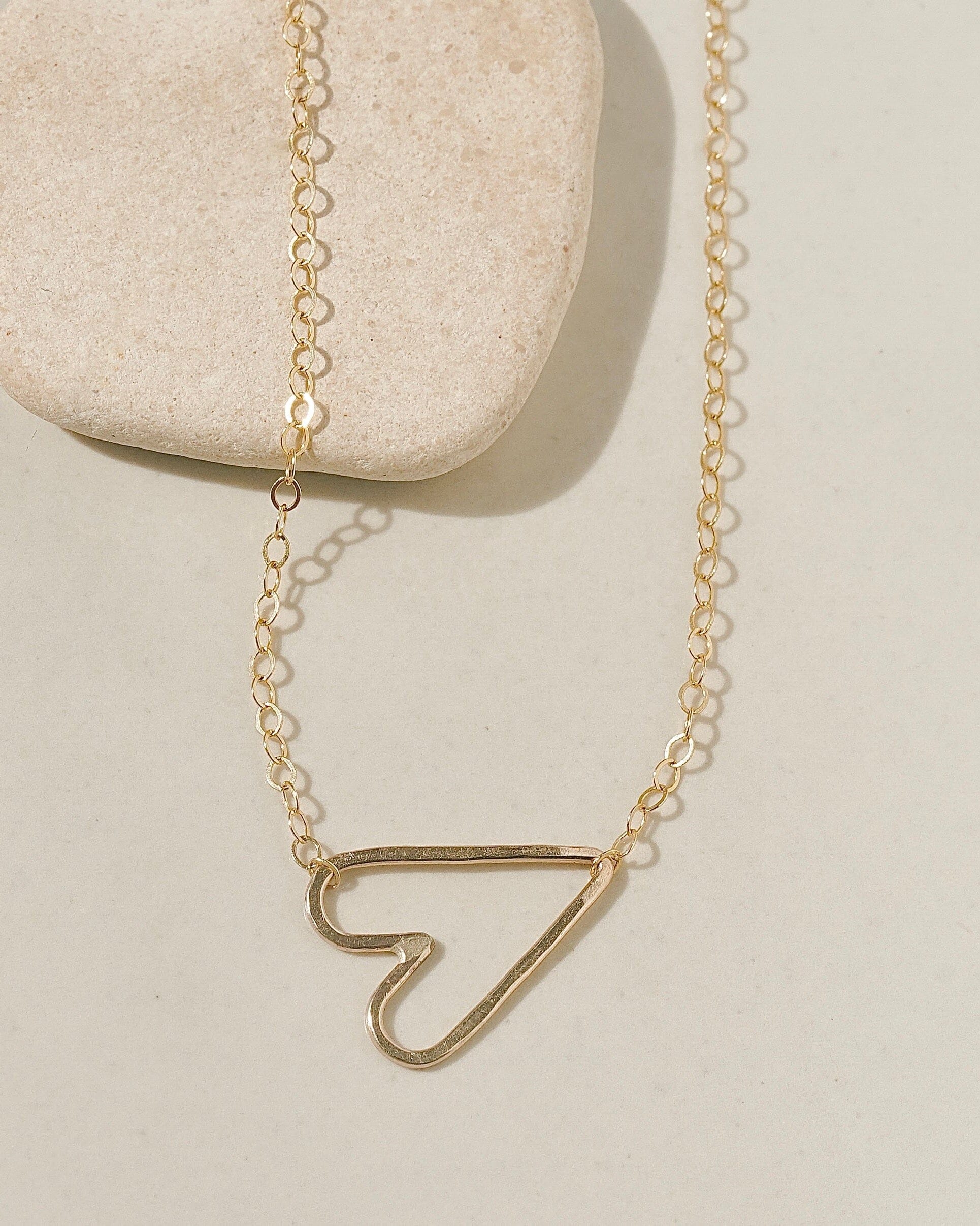 Delicate gold chain necklace with a sideways open-outline gold heart pendant on cream surface with stone pebble