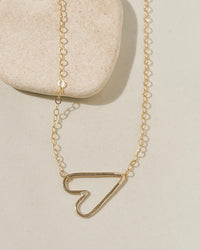 Delicate gold chain necklace with a sideways open-outline gold heart pendant on cream surface with stone pebble
